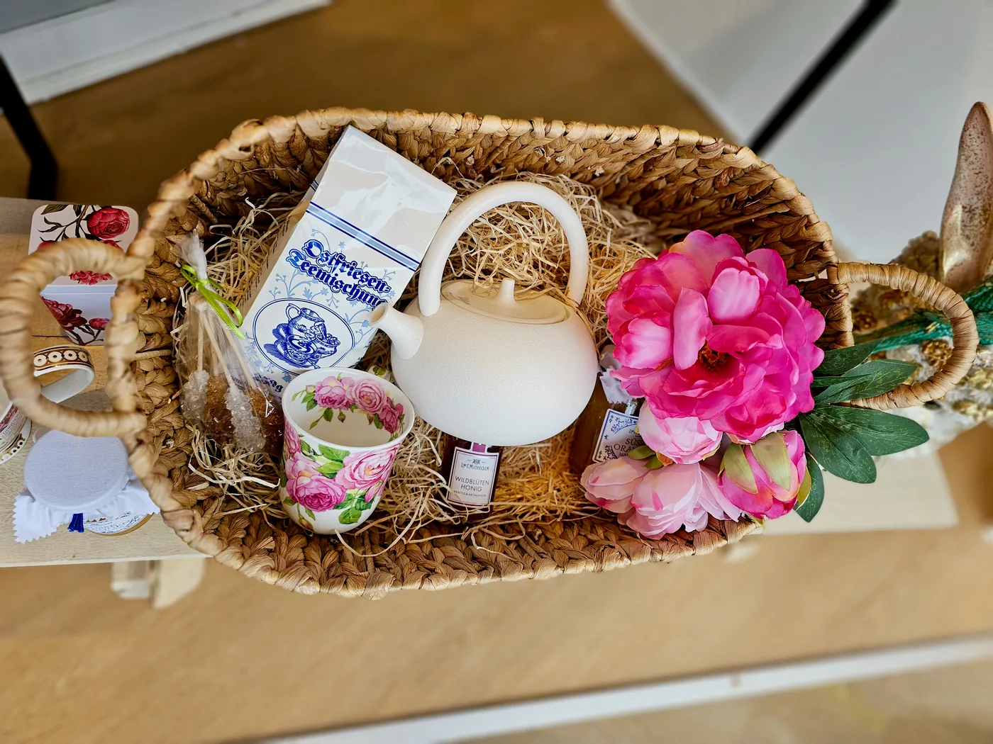 Ein geflochtener Geschenkkorb mit weißer Teekanne, Rosentasse, Honig und Tee, geschmückt mit rosa Blüten