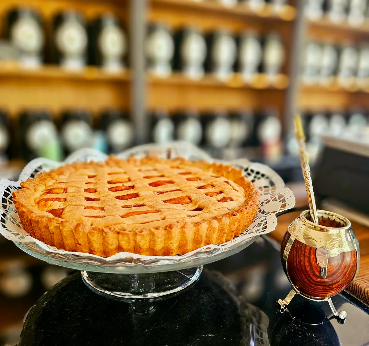 Ein goldbrauner Kuchen mit Teiggitter auf einer Glasplatte neben einem traditionellen Mategefäß mit Bombilla