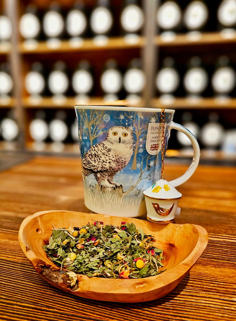 Blaue Dunoon-Porzellantasse mit Schneeeule und einem kleinen Teesieb neben einer Holzschale mit losem Kräutertee